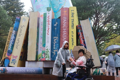 책모양 조형물앞에 휠체어에 앉아있는복지관이용자와 옆에 서있는 복지관이용자 사진
